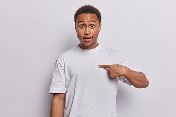 Photo of shocked young African man with dark curly hair and full lips points at himself with stunned expression holds breath wears casual t shirt isolated over white background. Do you mean me