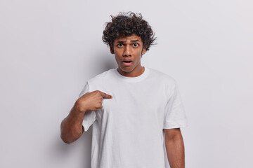 Waist up shot of confused curly haired Hindu man points at himself and has puzzled expression asks who me being stunned to be mentioned or chosen dressed in casual basic t shirt isolated on white wall