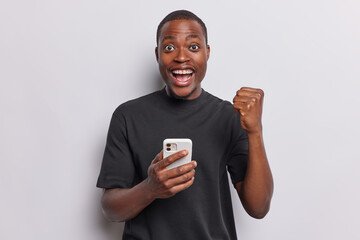 Cheerful dark skinned African man holds smartphone clenches fist and celebrates good news smiles...