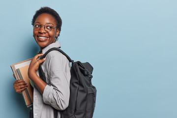 Horizontal shot of cheerful African woman stands sideways with backpack carrying notepads and...