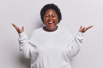 Waist up shot of dark skinned African woman keeps hands raised up exclaims loudly has eyes closed dressed in casual sweatshirt reacts to something isolated on white background. Human reactions concept - obrazy, fototapety, plakaty