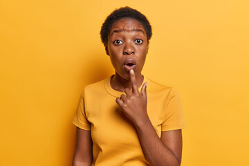 Omg concept. Stunned African girl with short hair feels amazed has widely opened mouth keeps finger on lips dressed in casual t shirt isolated over vivid yellow background emotionally reacts to news - obrazy, fototapety, plakaty