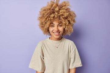 Indoor shot of cheerful curly haired millennial girl smiles happily dressed in casual t shirt looks directly at camera expresses positive emotions isolated over purple background. People and happiness