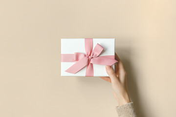 Female hand holding a white gift box with a pink bow on a beige background, top view. Mother's Day, Valentine's Day, Christmas and New Year gifts concept