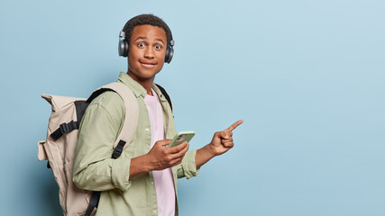 Horizontal shot of surprised dark skinned man holds mobile phone connected to headphones points...