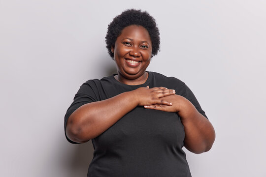 Waist up shot of cheerful overweightt African woman presses both hands to heart smiles gladfully expresses gratitude wears casual black t shirt isolated over white background. Body language concept