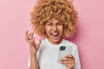 Annoyed curly haired woman holds smartphone gestures actively expresses negative emotions screams from anger wears casual white t shirt expresses negative emotions isolated over pink background