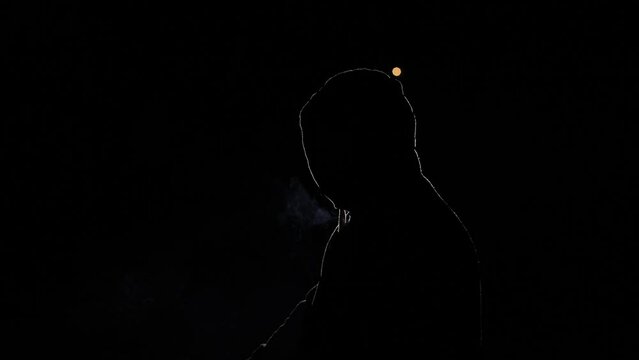 Silhouette of a person on a black background. A man in a hood, illuminated by light, blows. A hacker in the night, a mysterious person whose face is not visible. He smokes a cigarette