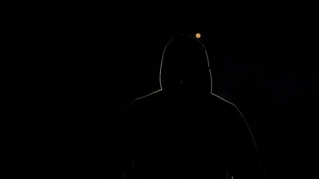 Silhouette of a person on a black background. A man in a hood, illuminated by light, blows. A hacker in the night, a mysterious person whose face is not visible. He smokes a cigarette