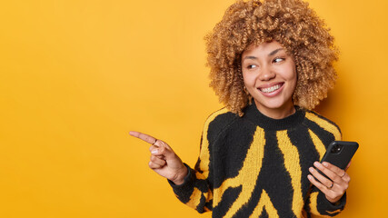 Horizontal shot of pretty curly haired young woman in casual black and yellow jumper holds mobile phone chats with friends shows blank space for your promotion poses indoors. Technology concept