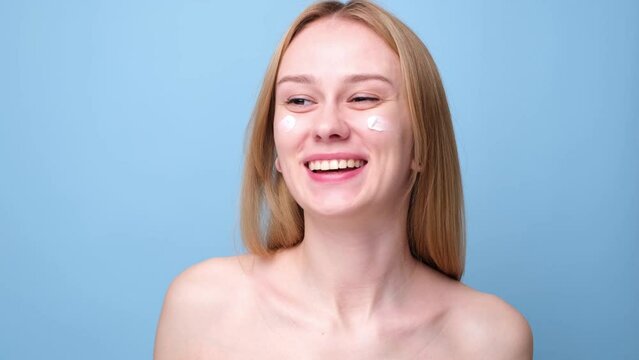A beautiful girl applies moisturizer to her facial skin. Young woman on blue background smiling and taking care of her skin.