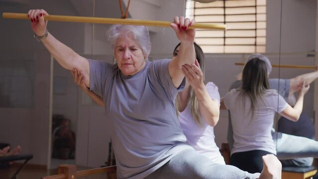 Pilates instructor guiding a senior woman in physiotherapy class with wooden stick and stretching leg