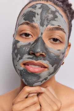 Headshot of serious Latin woman touches chin gently and looks directly at camera applies nourishing facial clay mask on face for skin treatment isolated over white background. Wellbeing concept