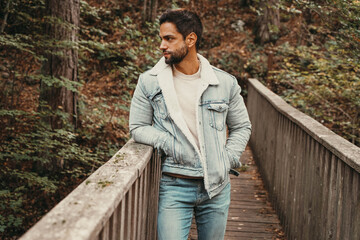 A charismatic young man showcasing style in a denim jacket and jeans, striking a confident pose on an outdoor wooden bridge