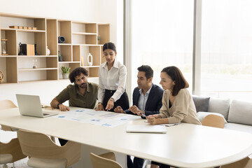 Serious young project manager woman explaining sales marketing colleagues, investors, standing at meeting table, pointing at document with statistic data, speaking to partners