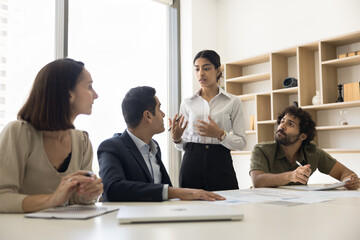 Serious young Indian professional woman coaching company staff, talking to business colleagues on cooperation meeting, explaining project plan, standing at table, speaking