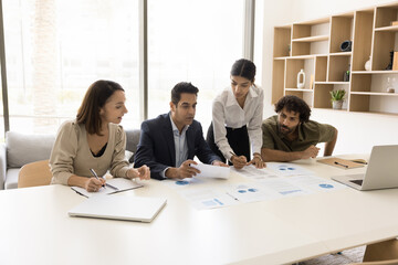 Serious Indian team leader woman presenting sales reports to team. Company coach giving marketing seminar to staff, diverse group of employees, explaining paper diagram on table
