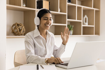 Cheerful young Indian manager woman in headphones online meeting, job communication on Internet, talking on video conference call, waving greeting hand hello at laptop