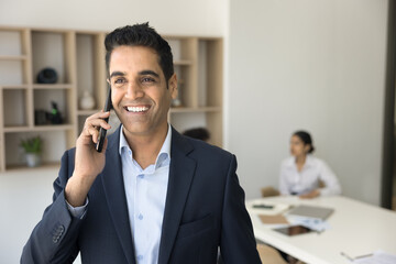 Cheerful successful handsome businessman enjoying mobile phone call, talking on cell, getting good news with toothy smile, laughing. Boss speaking on cell while team working in background
