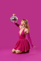 Young woman dressed as doll with disco ball sitting on purple background