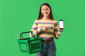 Young woman with shopping basket pointing at mobile phone on green background
