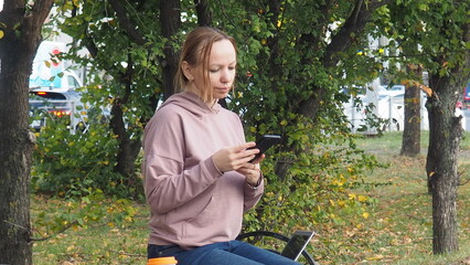 freelancer woman sit on bench in park, uses a smartphone, coffee to go, looks social networks, online training, conference. outdoors rest, talk by mobile cell phone . People urban lifestyle concept