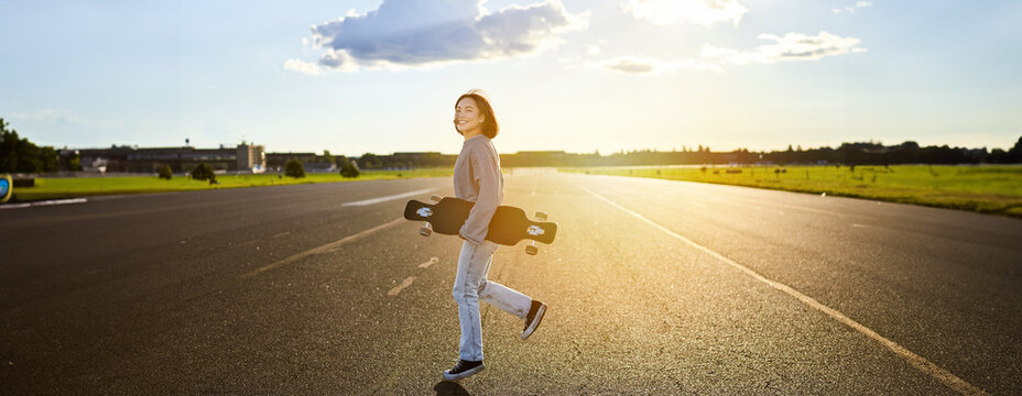 Asian girl with skateboard standing on road during sunset. Skater posing with her long board, cruiser deck during training