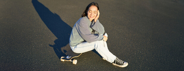 Portrait of asian woman sitting on skateboard, skating on her cruiser longboard, using smartphone...