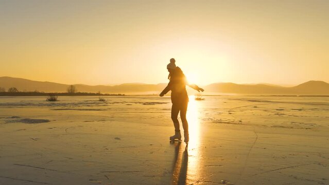 LENS FLARE, SILHOUETTE: A happy woman is skating on a golden shining frozen lake. She is having fun and enjoys dancing on vast empty natural ice rink as the winter sun is setting behind the hills.