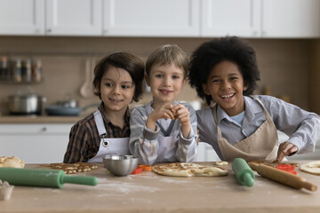 Fototapeta premium Three multiethnic preschoolers boys in aprons cooking together in kitchen, prepare surprise for family, make dough for homemade pizza posing smile look at camera at home. Cousins friendship and hobby