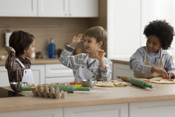 Three cute multiethnic little boys wear aprons give high five, cooking pizza, Italian cuisine...