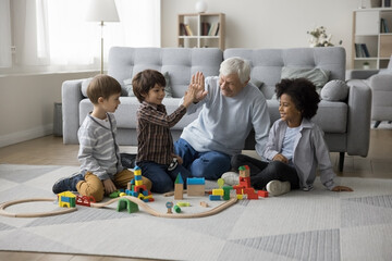 Fototapeta premium Great-grandpa gives high five to little great-grandson sit on floor spend pastime together with multiethnic great-grandchildren play wooden railroad, multi-generational family ties, games, playtime