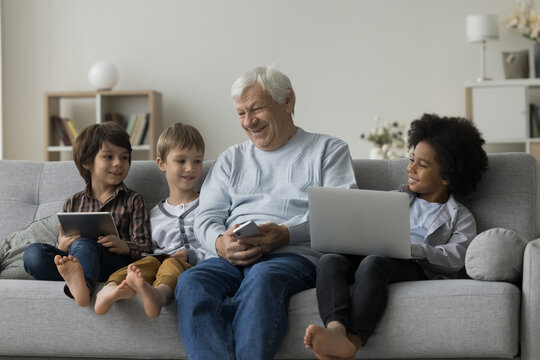 Loving older great-grandpa and great-grandsons sit on couch holding diverse modern devices, laugh, enjoy pastime on internet and communication. Modern technologies overuse, bad habit, leisure online