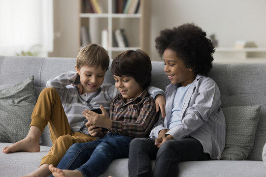 Three smiling cute boys sit on sofa in living room use mobile phone play online games on smartphone, watch funny video, friends spend free time together at home chatting with mates. Modern technology