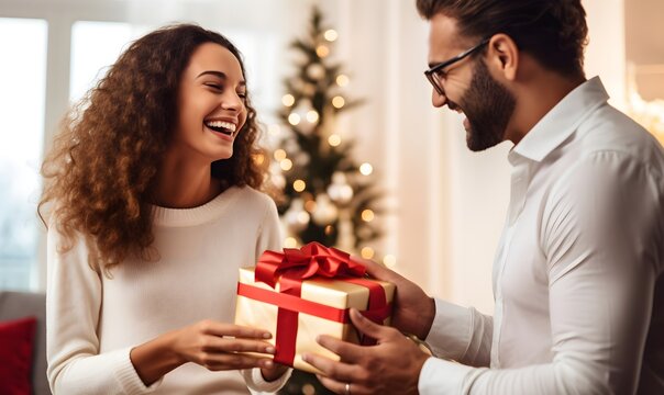 Happy couple exchanging Christmas gift with red bow on background of christmas tree with lights. Stylish multiethnic lovers holding present close up in festive decorated room. Happy Holidays, at home - Powered by Adobe