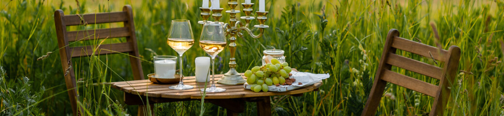 Beautiful romantic outdoor wedding decor in a field. Table decorated with burning candles, flowers. Wineglasses with white wine. Sunset, summer, golden hour. Perfect surprise date for couple. Banner