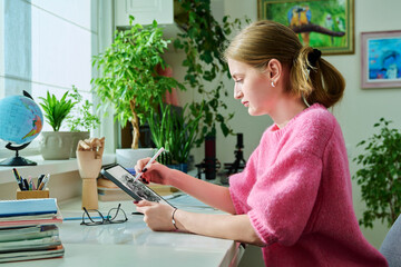 Teenage girl student illustrator drawing on digital tablet sitting at desk at home