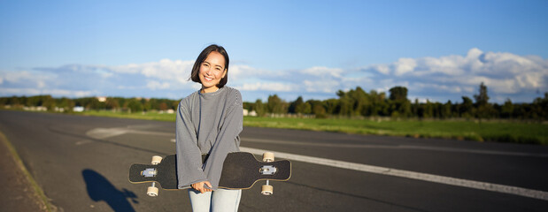 Vertical shot of skater girl posing with longboard, cruising on empty road in suburbs. Smiling...