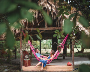 casal jovem deitado em rede cor de rosa em frente a uma maloca com teto de palha rodeado de árvores verdes, em Soure, ilha do Marajó  - obrazy, fototapety, plakaty