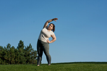 overweight middle-aged woman does exercises in park standing on grass, blue sky background. fat woman tilts body to side. Healthy lifestyle, body positivity, Pilates outdoors in summer