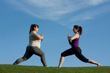 Two middle-aged women practice yoga in city park in warrior pose, blue sky background. Namaste. Healthy lifestyle, fitness, Pilates, weight loss. overweight woman doing yoga with trainer.