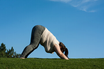 overweight middle-aged woman practices yoga in park, blue sky background. fat woman doing Downward...