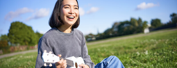 Happy people and hobbies. Smiling asian girl playing ukulele guitar and singing, sitting in park outdoors on blanket - Powered by Adobe