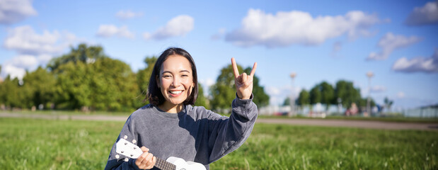 Happy asian girl playing ukulele in park, showing rock n roll, heavy metal horns sign and smiling,...