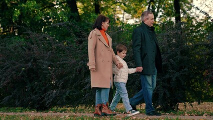 Naklejka na ściany i meble Happy child holding hands with mom dad walks through autumn park. Healthy family walk, mother, child, father in park. Kid son, mom, dad walk together holding hands in autumn outdoor. Mother father boy