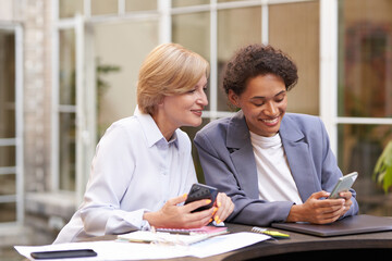 Smiling business colleagues discuss biz issue while use mobile phones sitting in office terrace