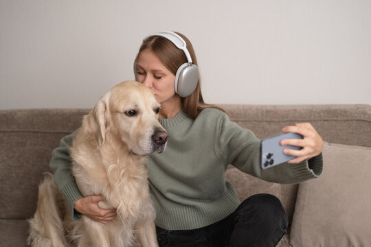 girl sitting with her dog on the couch with headphones on. A young woman takes a picture with her dog at home.