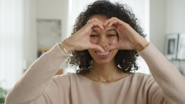 Smiling model in casual beige sweater posing showing LOVE. Portrait of attractive female student with curly brunette afro hair making heart shape with hands and looking to camera in modern apartment