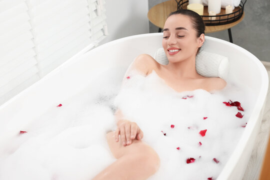 Happy woman taking bath in tub with foam and rose petals indoors