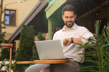 Handsome young man with laptop checking time on watch at table in outdoor cafe. Space for text